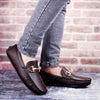Brown loafers with gold buckles worn with gray jeans against a brick wall background